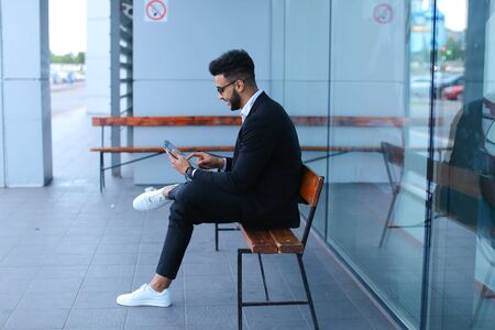 Entrepreneur smiling man puts on sunglasses uses tablet and looks at. Young handsome businessman arab muslims in business center wearing dressed in black elegant suit on building background.の写真素材