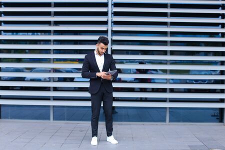 Young handsome businessman Arab Muslims is in the business center in black elegant dress on building background. Businessman man male looking waiting concept and smiling.の写真素材