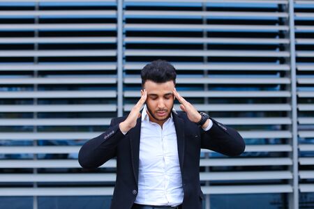 Entrepreneur shows neck pain tired looking at the camera. Young handsome businessman arab muslims in business center wearing dressed in black elegant suit on building background.の写真素材