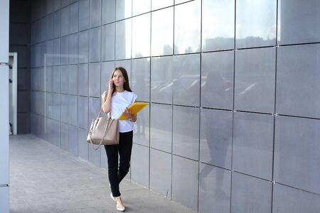 Successful young business woman walks and talks on phone, holds documents in yellow folder, smiling on background of gray wall of shopping business center.の写真素材