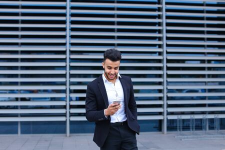 Entrepreneur uses phone mobile smart looking at the camera. Young handsome businessman arab muslims in business center wearing dressed in black elegant suit on building background.の写真素材