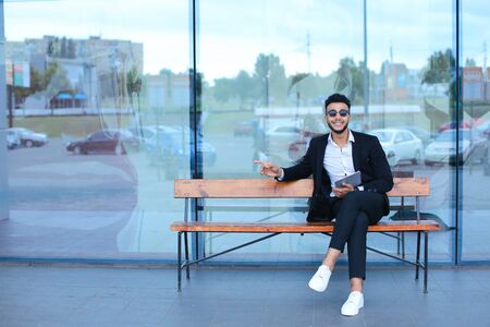 Entrepreneur smiling man puts on sunglasses uses tablet and looks at. Young handsome businessman arab muslims in business center wearing dressed in black elegant suit on building background.の写真素材