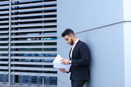 Perspective young adult businessman virile asian guy holds and looks, considers, reads and sorts out papers or documents, stands near gray wall of business center.の写真素材
