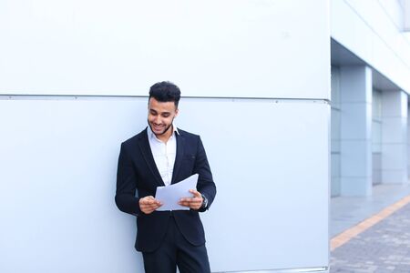Young successful student businessman arab man holds documents, securities, smiling and thinks near gray wall of business center.の写真素材