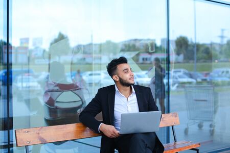 Entrepreneur uses laptop wearing sunglasses. Young handsome businessman arab muslims in business center wearing dressed in black elegant suit on building background.の写真素材