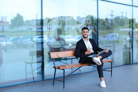 Entrepreneur uses laptop wearing sunglasses. Young handsome businessman arab muslims in business center wearing dressed in black elegant suit on building background.の写真素材