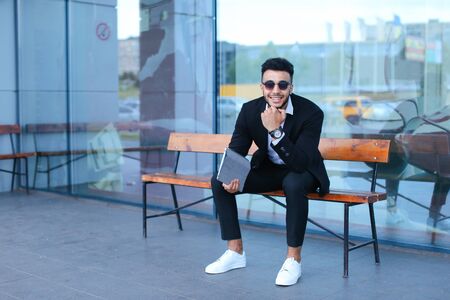 Entrepreneur smiling man puts on sunglasses uses tablet and looks at. Young handsome businessman arab muslims in business center wearing dressed in black elegant suit on building background.の写真素材