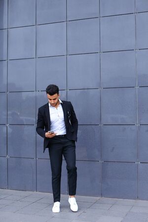 Entrepreneur man male looking at camera and smiling talks on mobile smart phone. Young handsome businessman arab muslims in business center wearing dressed in black elegant suit on building background.の写真素材
