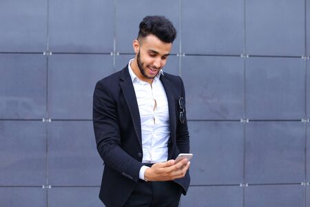 Entrepreneur takes makes selfie man male looking at camera and smiling talks on mobile smart phone. Young handsome businessman arab muslims in business center wearing dressed in black elegant suit on building background.の写真素材
