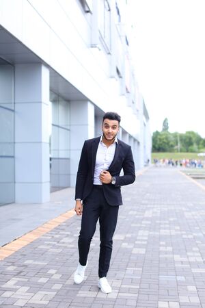 Young handsome businessman Arab Muslims is in the business center in black elegant dress on building background. Businessman man male looking at time walking goes and smiling.の写真素材