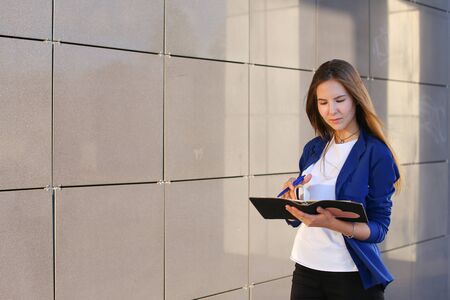 Pretty female girl student holds in hands diary, notebook, Students work, homework and writes pen, constitutes plan against backdrop of modern business center.の写真素材