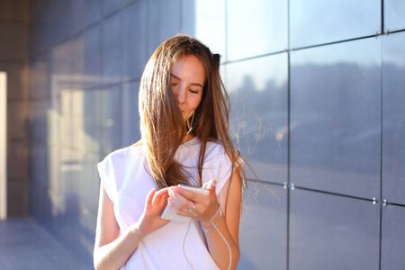 Nice young female girl business woman student holds white telephone, listening and enjoying music in headphones, stands near wall of modern business center. Girl with long light brown hair dressed in white shirt. Concept of headphone advertisement, mobile phone, technology, listening of good music, student everyday life.の写真素材
