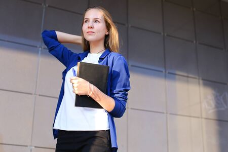 Young student beautiful girl female woman confidently goes forward, striding to dream and holds notebook, diary and pen in hand on background of business center outdoors.の写真素材