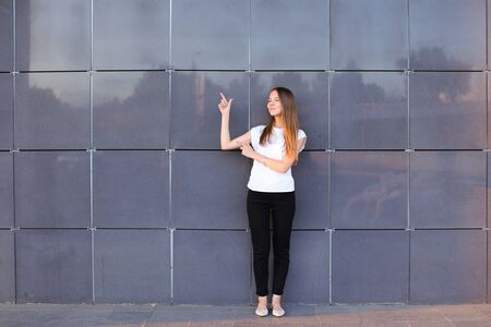 Beautiful young energetic female girl raised arms and points to advertising space, worth exactly in full growth and smiles on background of gray wall of modern business center.の写真素材