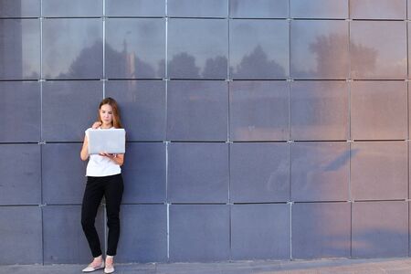 Promising young entrepreneur beautiful woman uses laptop for work, make appointment, solves important business issues and and stands right in all growth against the background of gray wall of modern business center.の写真素材