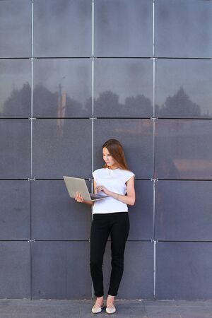 Promising young entrepreneur beautiful woman uses laptop for work, make appointment, solves important business issues and and stands right in all growth against the background of gray wall of modern business center.の写真素材