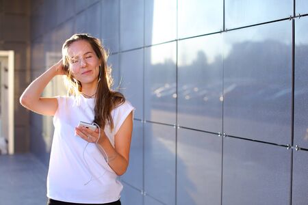 Nice young female girl business woman student holds white telephone, listening and enjoying music in headphones, stands near wall of modern business center. fe.の写真素材