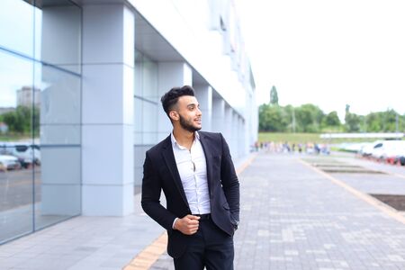 Young handsome businessman Arab Muslims is in the business center in black elegant dress on building background. Businessman man male looking at time walking goes and smiling.の写真素材