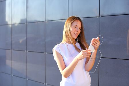 Beautiful cheerful young female student girl holding white phone, smiling, laughs and chatters on headphones.の写真素材