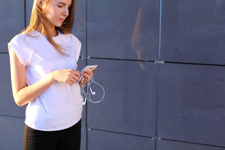 Lovely girl young female woman unravels white headphones for listening to music, holds in hands phone, smiles and stands near gray wall of modern business center.の写真素材