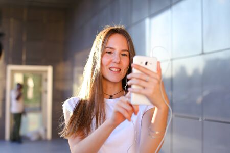 Beautiful cheerful young female student girl holding white phone, smiling, laughs and chatters with headphones.の写真素材