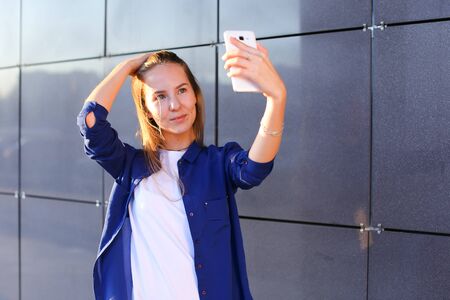 Charming cute lady girl student photographed, makes selfie photos and smiling at camera on background of business center on outdoors. Girl with long light brown hair dressed in white T-shirt and blue shirt. Concept of Concept of takes selfie, cellphone, makes phoro, holidays, student weekdays, technology.の写真素材