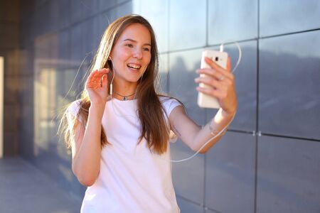 Beautiful cheerful young female student girl holding white phone, smiling, laughs and chatters on Skype with headphones, waves hand, welcomes or says goodbye. Girl with long light brown hair dressed in white shirt. Concept of headphone advertisement, mobile phone, Skype, technology, listening of good music, student everyday life.の写真素材