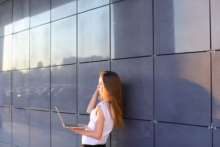 Beautiful young female woman girl entrepreneur successful person holds laptop in hands works, solves problems, and stands sideways to camera on background wall of modern business center.Girl with long hait dressed in white shirt and black trousers.Concept of business meeting, business negotiations, advanced technology, gadget.の写真素材