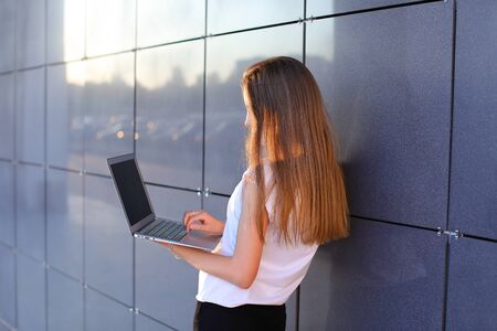 Young successful woman entrepreneur girl keeps in hands computer and works, typesets text, resolves issues stands sideways to camera on background of wall of business center. Girl with long hait dressed in white shirt and black trousers.Concept of business meeting, business negotiations, advanced technology, gadget.の写真素材