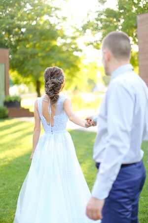Caucasian groom following bride and walking in park holding hands. concept of wedding and bridal photo session in open air.の写真素材