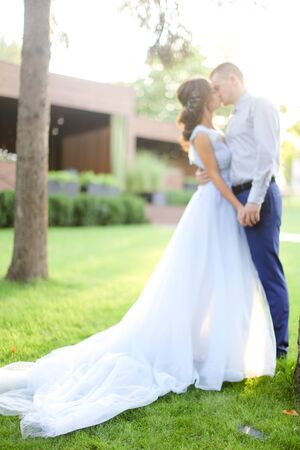 Caucasian fiancee kissing groom in park and wearng white dress. Concept of married couple and bridal photo session in open air.の写真素材