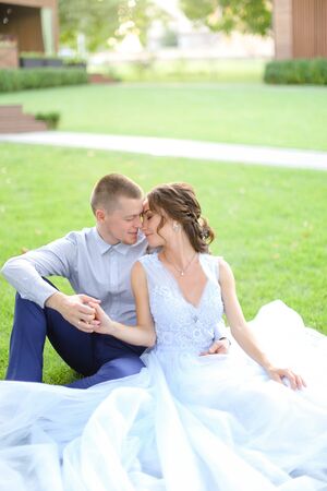 Caucasian pretty bride with groom sitting and kissing on grass in park, wearing white amazing dress. Concept of bridal photo session in open air, beautiful fiancee.の写真素材