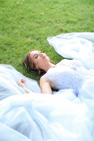 Innocent beautiful fiancee lying on grass in oark and wearing white dress. Concept of beauty and bridal photo session in open air.の写真素材