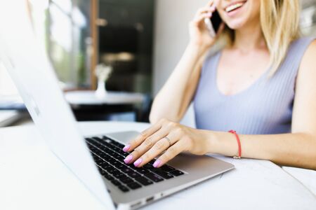 Blonde woman talking by smartphone and surfing internet by laptop at cafe. Concept of online shop assistant.の写真素材