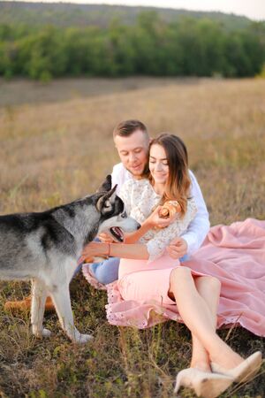 Young man and girl sitting on pink plaid outside and playing with malamute. Concept of pets and picnic on nature, happy couple and dog.の写真素材