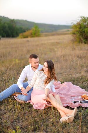 Young wife and husband sitting on pink plaid in field. Concept of picnic in open air and relationship, love.の写真素材