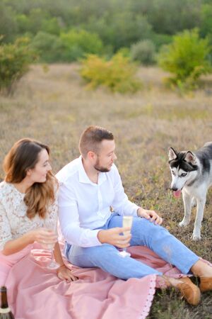 Young man and woman sitting on pink plaid with glasses near dog. Concept of picnic on nature and pets.の写真素材