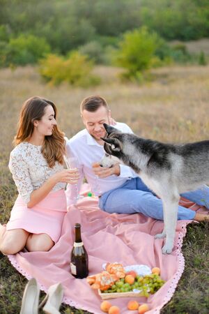 Young girl and man drinking wine on pink plaid near dog. Concept of resting on nature, picnic and pets.の写真素材