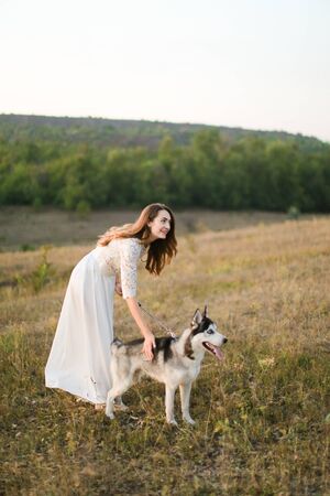 Caucasian nice woman walking in steppe with dog husky and wearing white dress. Concept of nature, bridal photo session on nature and beauty, pets.の写真素材