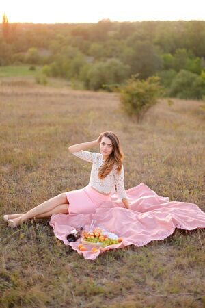 Young pretty girl sitting on pink plaid near bottle of red wine and fruits. Concept of resting in open air and summer picnic.の写真素材