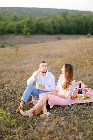 Young boy blowing soap bubbles near caucasian girl and sitting on pink plaid in steppe. Concept of happy couple, romantic love and picnic on nature.の写真素材