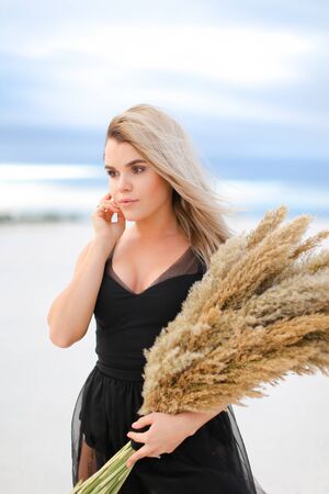 Young blonde woman wearing black dress standing with dried plants bouquet. Concept of fashion and seasonal winter photo session.の写真素材