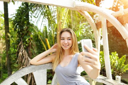 Young woman taking photo selfie on smart phone on sunny day, background of sunshine green palms in Thailand, Phuket. Concept of social networks, new technologies for better life, travel to tropical countriesの写真素材
