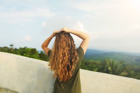 Back view of young girl with raised hands wearing khaki dress, standing in mountains background. Concept of summer vacations and resting on nature.の写真素材