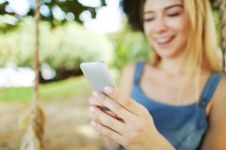 Focus on smartphone hold by young blonde woman riding swing and smiling, Thailand. Concept of modern technology, internet and summer vacations in tropics.の写真素材