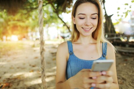 Sunshine portrait of young blonde girl chatting by smartphone and riding swing in Bali, sand in background. Concept of modern technology, youth and summer vacations.の写真素材