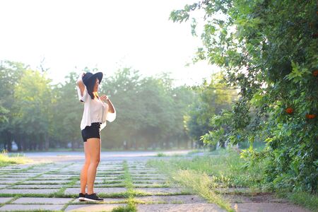 female nature keeps his hat summer sun sunset looks pleased. azitka woman black hair white shirt black shorts dancing around the trees looking at the camera. portrait. spinningの写真素材