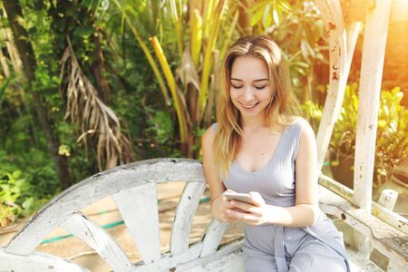 Young happy woman uses smartphone for work on sunny day, background of sunshine green palms in Thailand, Phuket. Concept of new technologies for better life, travel to tropical countries, remote work freelancingの写真素材