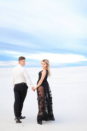 Young blonde girl in black transparent dress holding man hands and walking in winter steppe. Concept of seasonal photo session and love.の写真素材