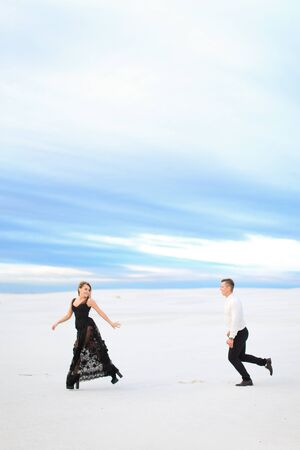 Young woman and man running on snow, wearing black transparent dress. Concept of winter photo session and happy couple.の写真素材
